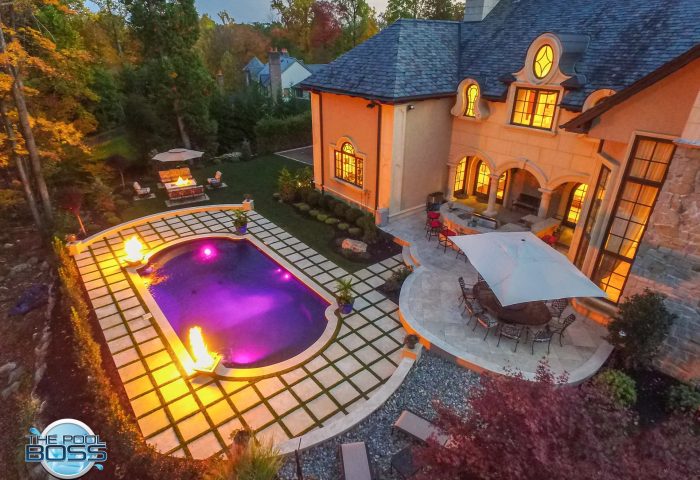 A luxurious backyard features a rectangular pool with glowing purple lights and two fire features. Theres a large patio with tables, chairs, and umbrellas beside a grand house at dusk, surrounded by trees and landscaping.