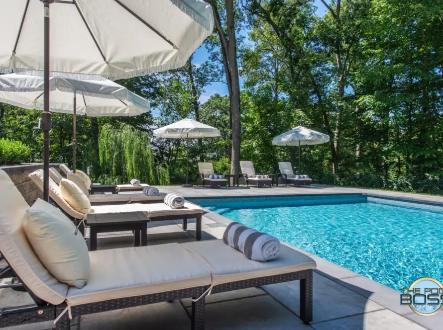 A luxurious outdoor pool area with lounge chairs, each topped with cushions and rolled towels, sits beside turquoise water. Large white umbrellas provide shade, surrounded by green trees.