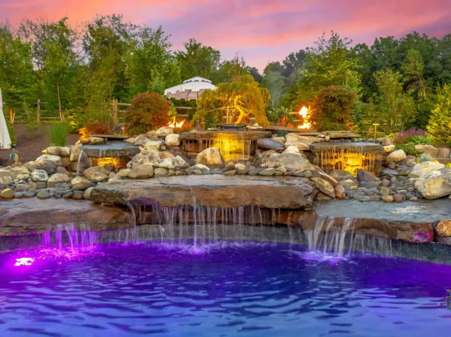 A landscaped backyard pool with purple underwater lights, a small waterfall over rocks, and fire features at sunset. Outdoor seating and lush greenery surround the area. The Pool Boss logo appears in the corner.